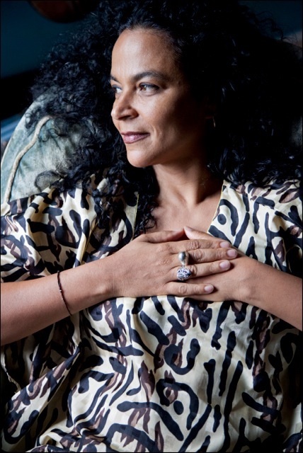 Gabri Christa wears a white and purple printed blouse. She sits in chair and we see her profile. Her hands rest on her chest.